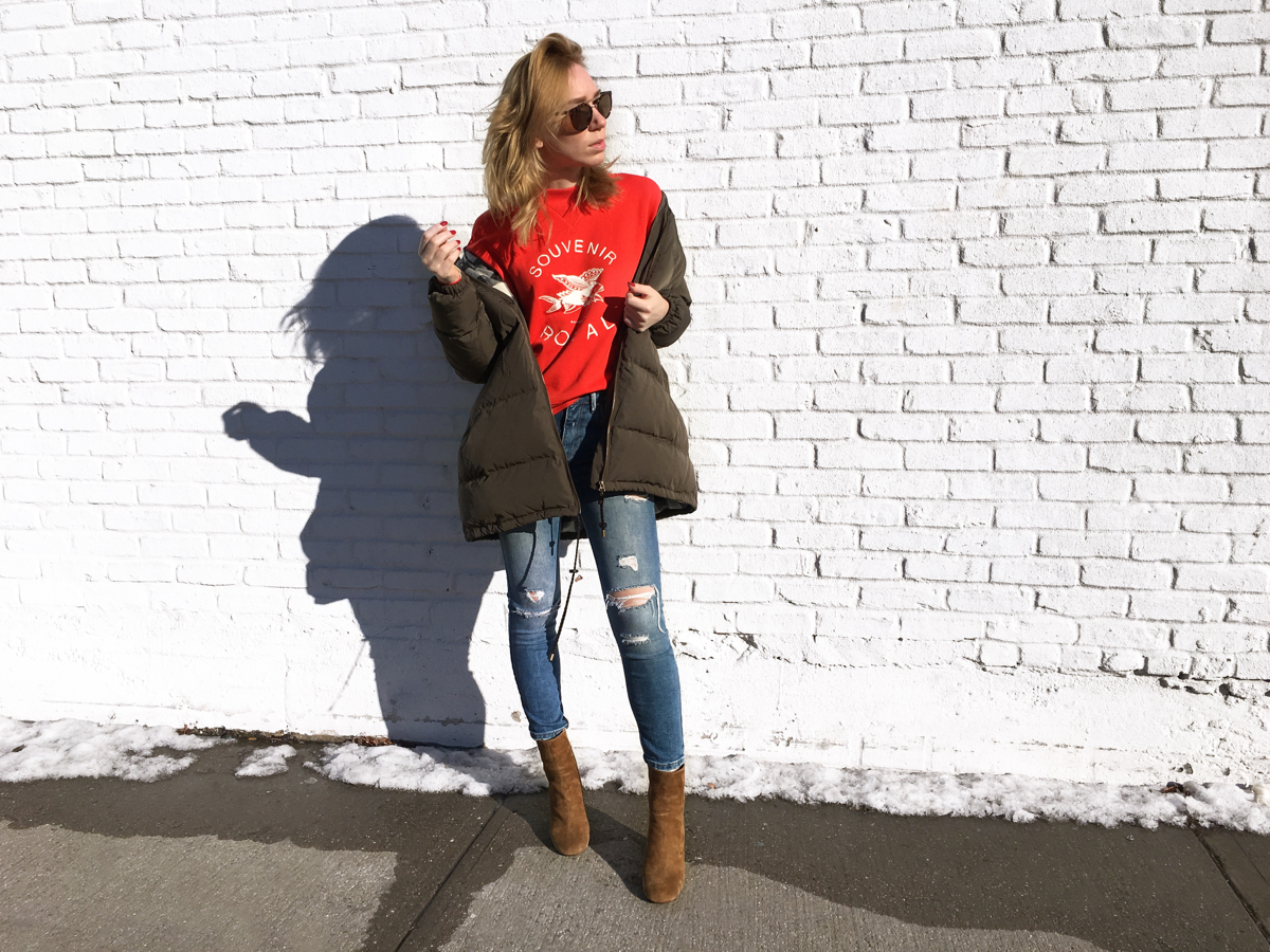 Woman posing against white brick wall wearing red sweater and olive jacket