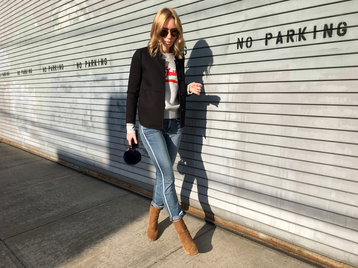 Woman walking on sidewalk wearing denim and grey sweatshirt outfit with navy coat
