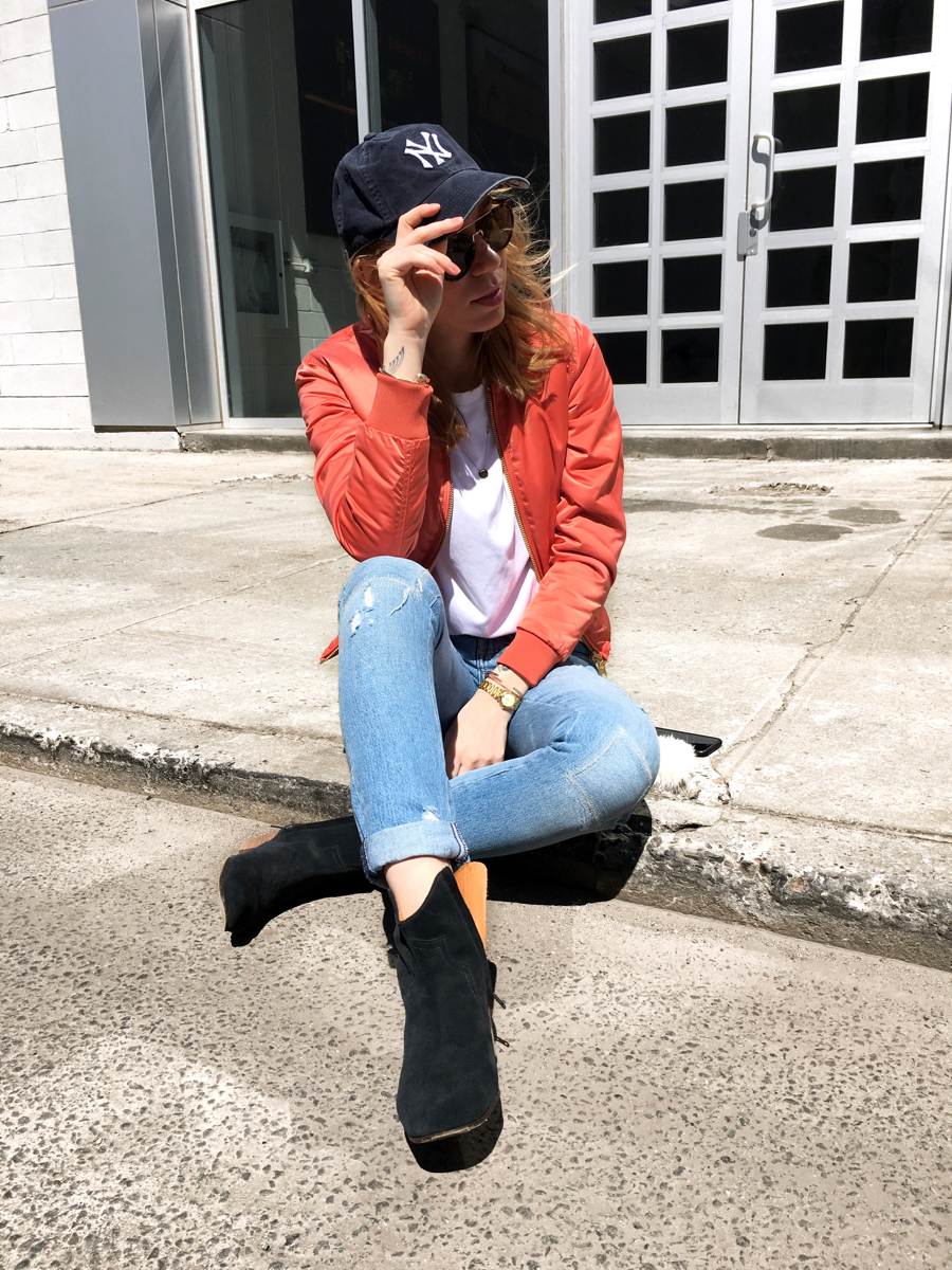 Woman sitting on sidewalk wearing pink bomber with Yankees cap