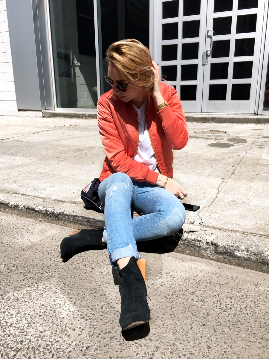 Woman sitting on sidewalk wearing pink bomber with jeans